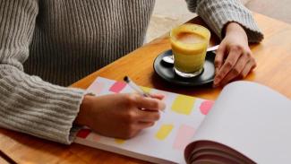 Person writing in notepad with coffee on desk.